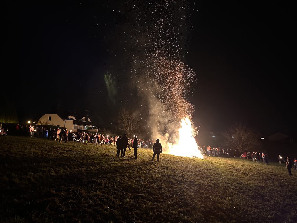 Auf einer Wiese brennt ein großes Feuer und erleuchtet den Nachthimmel mit Funkenflug. Viele Menschen stehen in sicherem Abstand drum herum, die Kinder halten bunte Laternen in der Hand.